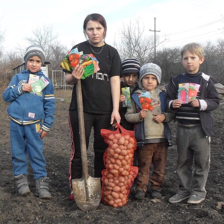 Mutter mit 4 Kinder stehen auf einem Feld und halten eine Schaufel, diverse Gemüsesaatcouverts und einen Sack kleine Kartoffeln
