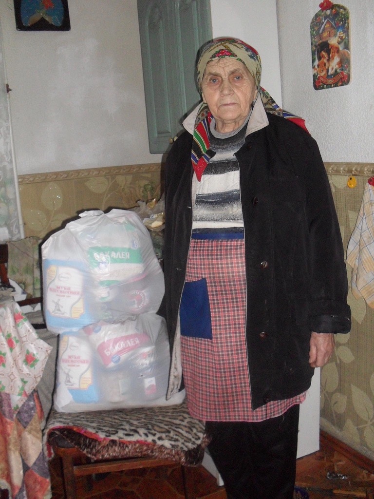 Eine alte Frau steht neben zwei Stopp-Hunger-Paketen - 2 weisse Säcke mit Nahrungsmittel darin
